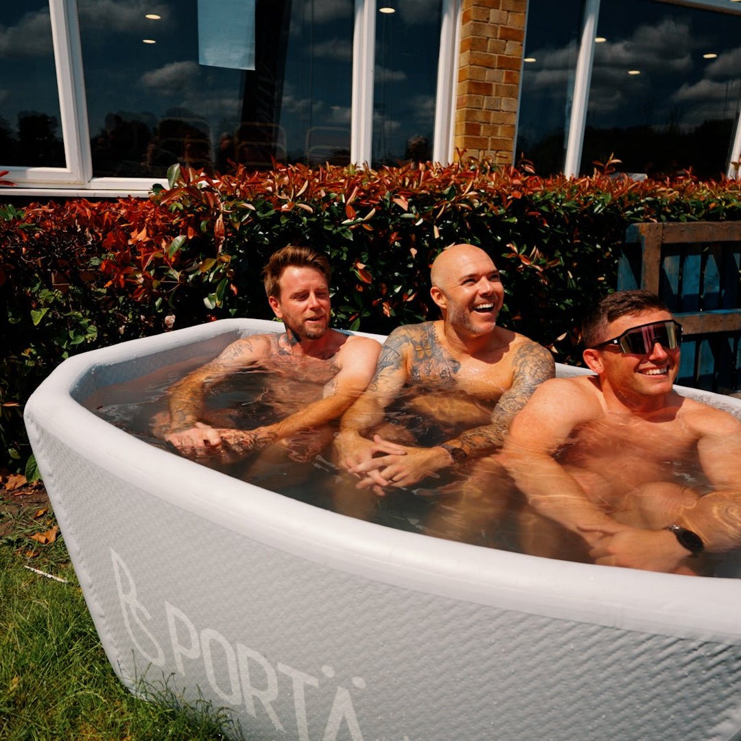 Three men in a portable hot tub outdoors with a brick building and plants in the background.