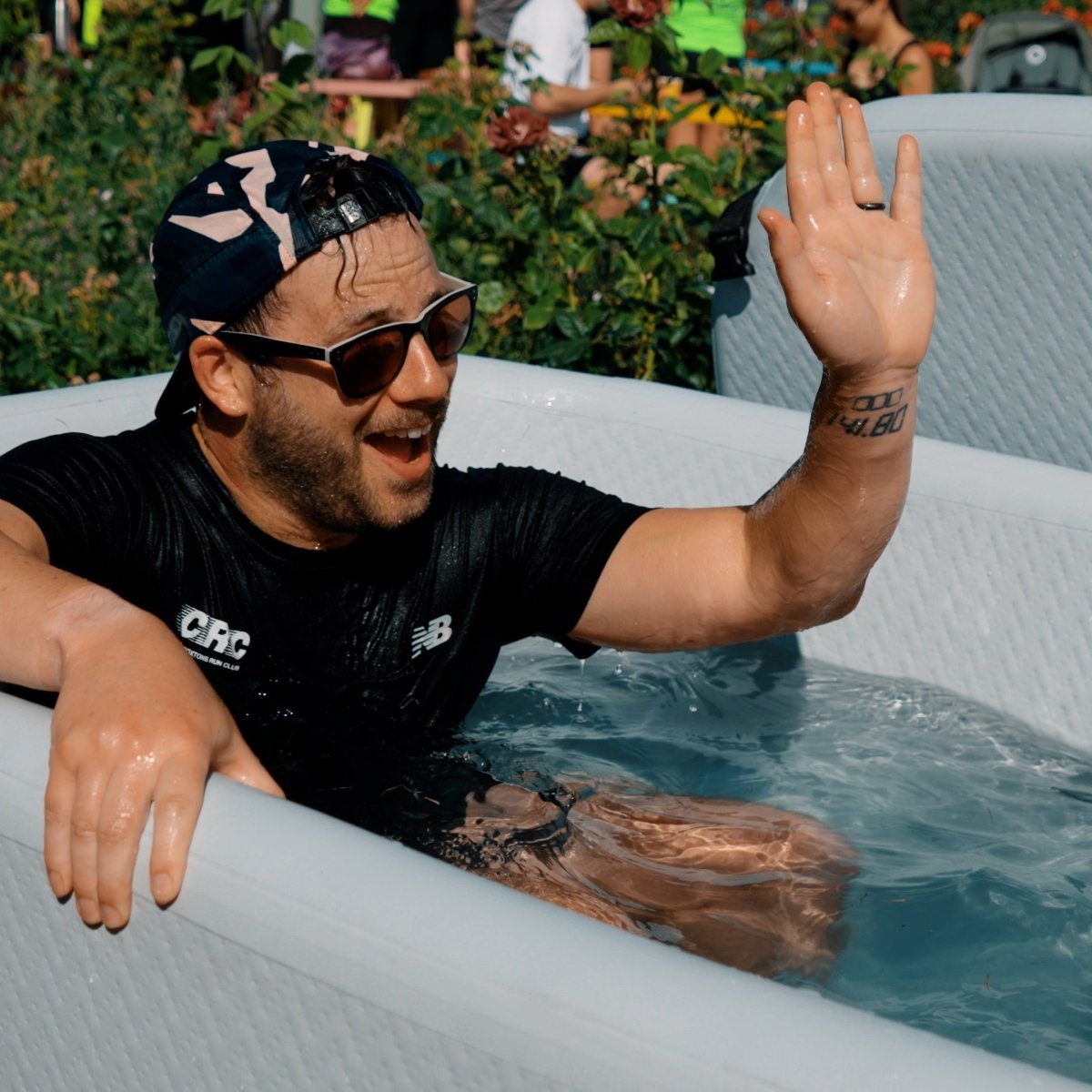 Man in a black shirt and sunglasses in a hot tub with another person reaching out to high-five.