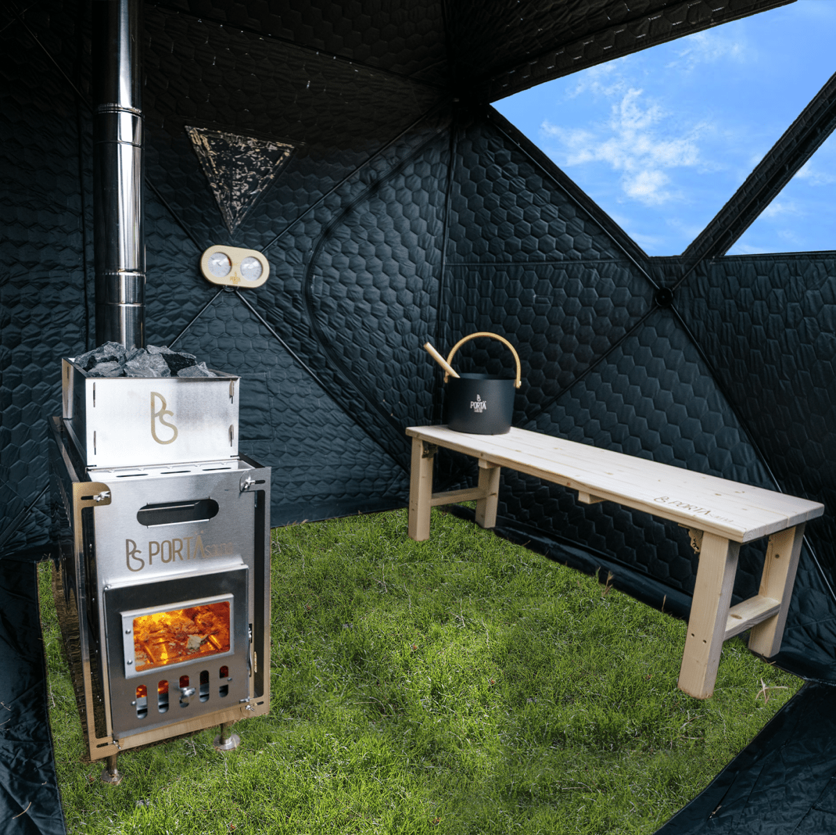 A portable wood-burning stove with a chimney stack sits on grass inside a black camping tent, next to a wooden bench and a black kettle. The tent interior is visible, with a blue sky seen through the window.